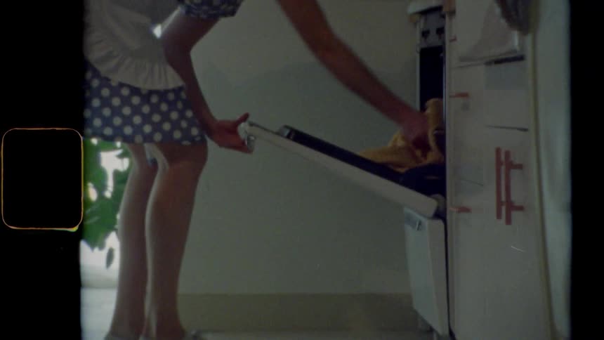 A woman wearing a dress and apron leans over to take a tray of food out of the oven
