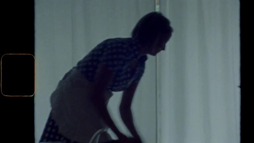A woman in an apron vacuums a rug in a dimly lit home kitchen during the day