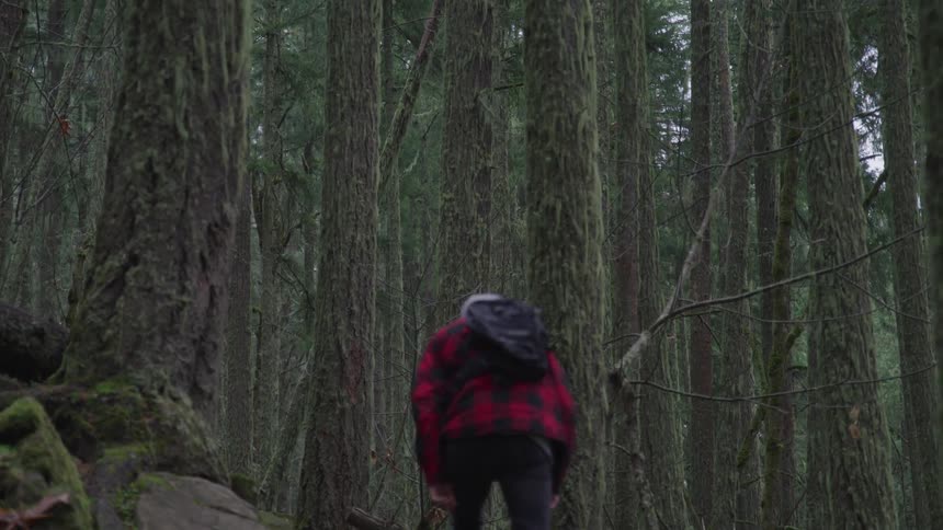 Person with white hair climbs sloping area in forest wearing backpack and plaid coat