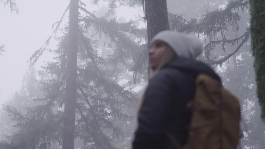 Woman with a with a white beanie a black coat and a brown backpack looks to the left in the foggy forest