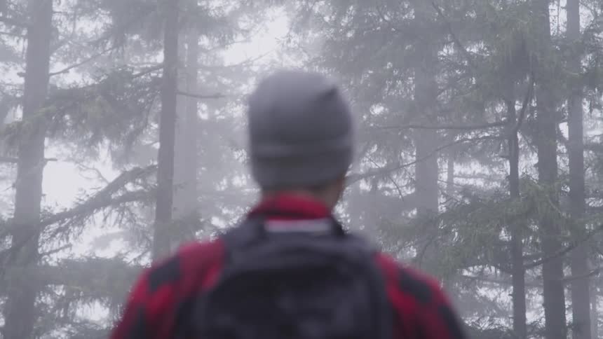 Man with a grey beanie a red plaid shirt and a black backpack looks around in the foggy forest