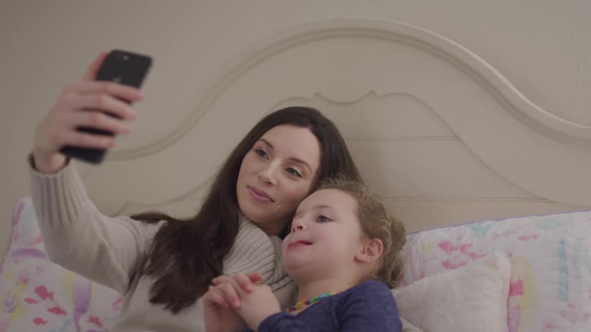 A mother and child lay in a bed holding a cell phone up for a selfie