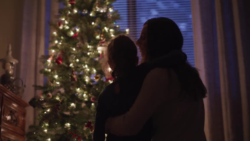 A woman hugs and kisses her daughter on the forehead as they gaze at the lights on a christmas tree together