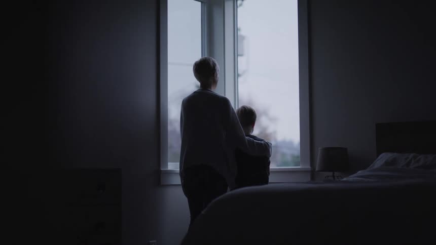 Mother with a short blonde hair holds her son and they look out the window in bedroom