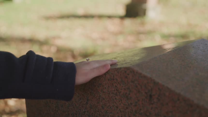 A woman runs her hand along the top of a headstone at a cemetery