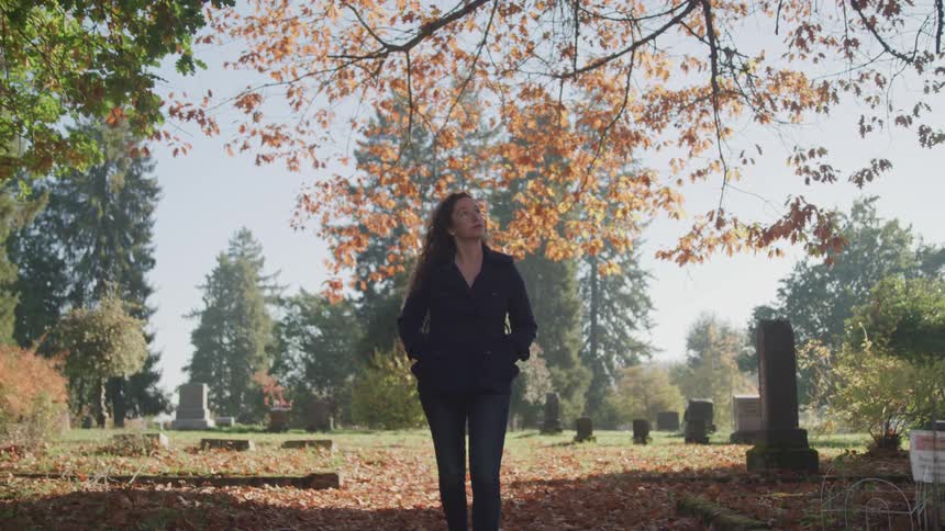 A woman walks through a cemetery with fall leaves all over the ground