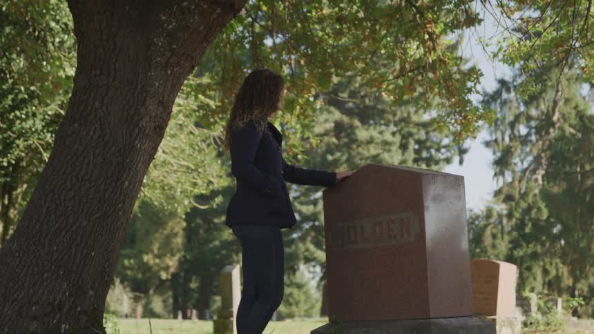 Woman stands meditatively in front of a headstone under a tree in the cemetery