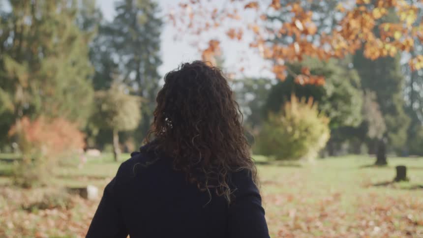 Woman with curly hair and a black overcoat walks on brown leaves in public park