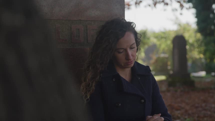 A white woman with long curly brown hair in a jacket is leaning against a large gravestone in the middle of a leaf filled cemetery with a sad look on her face