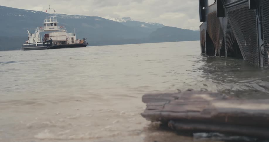 Ferry crosses ocean toward snow capped mountains as waves hit shore near dock