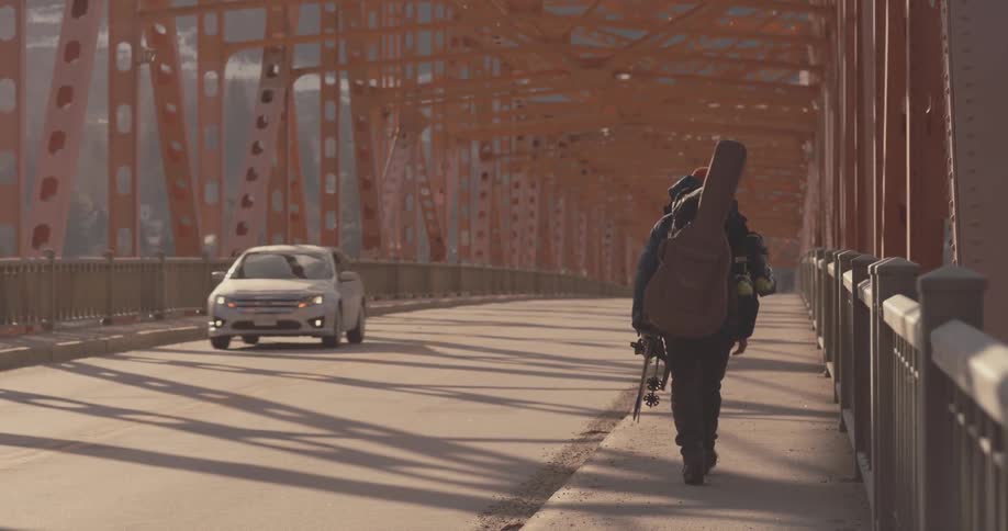 A man with a large guitar case strapped to his back walks along a sidewalk on a bridge