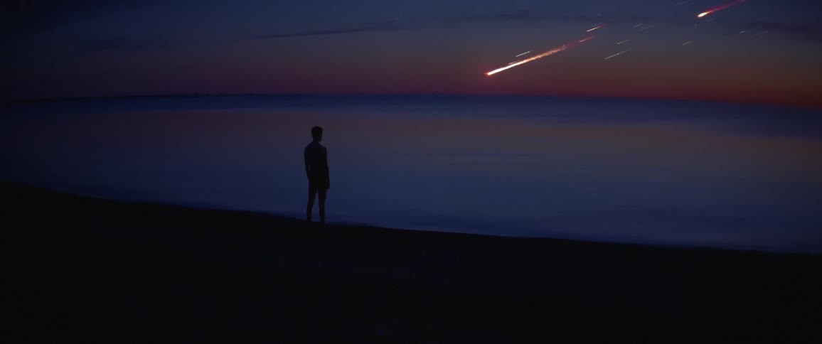 A man stands on a dark beach watching a shower of meteors on the horizon