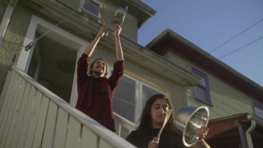A man and woman stand outside their home and bang on metal bowls with spoons