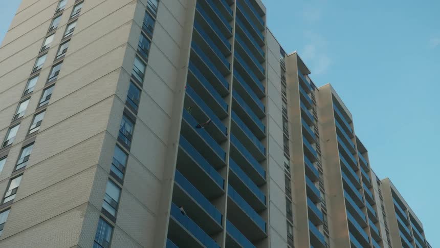 A woman claps on the side of her balcony while a few floors up another person claps two items together to the beat