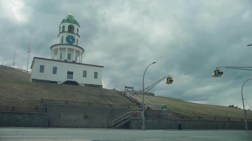 A single car travel on a road past a building with a bell tower and clock on a cloudy day