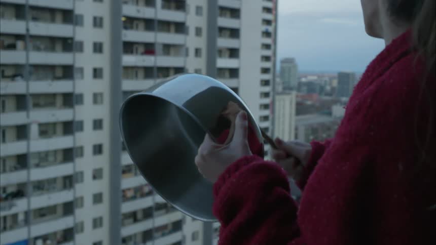 A woman bangs a pot with a wooden spoon while standing on an apartment balcony in a city