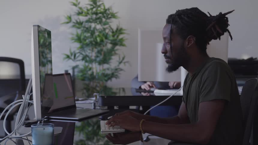 A black man sits at a desk while using a computer in an open office
