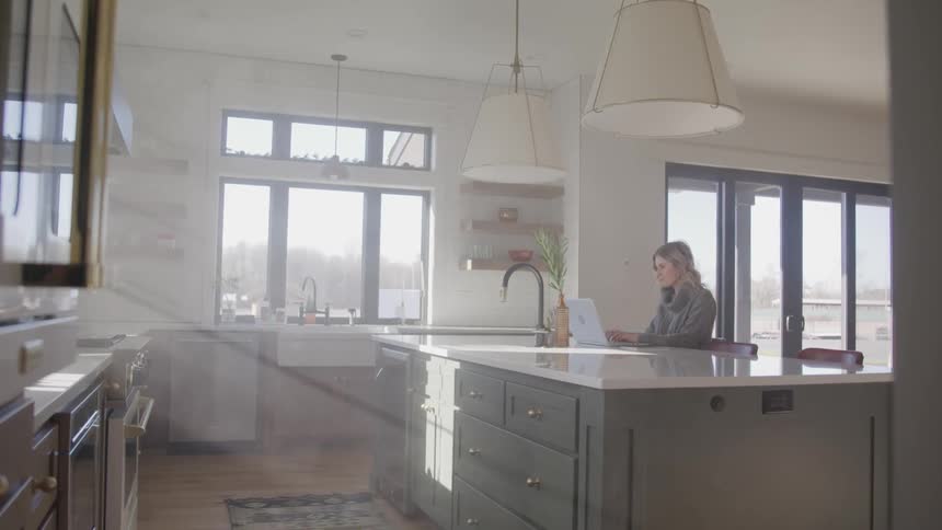 Woman sits at kitchen island in shadowy kitchen typing on laptop