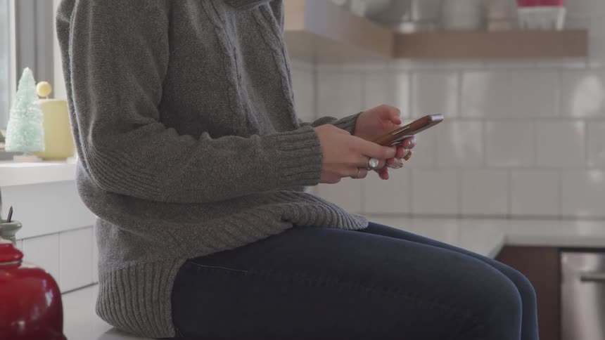 Woman sitting on the kitchen counter top texting on her smartphone