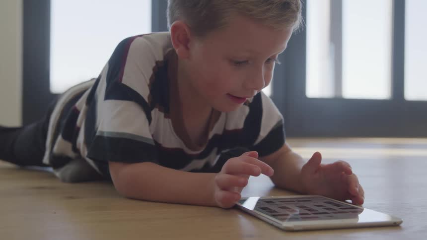 Blonde boy lies on the floor and uses a tablet at home