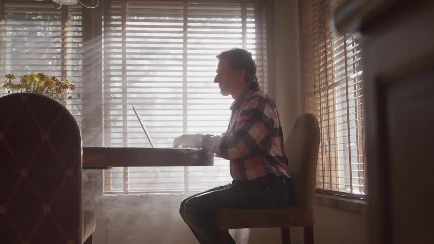 A man sits at a dining table typing on a laptop