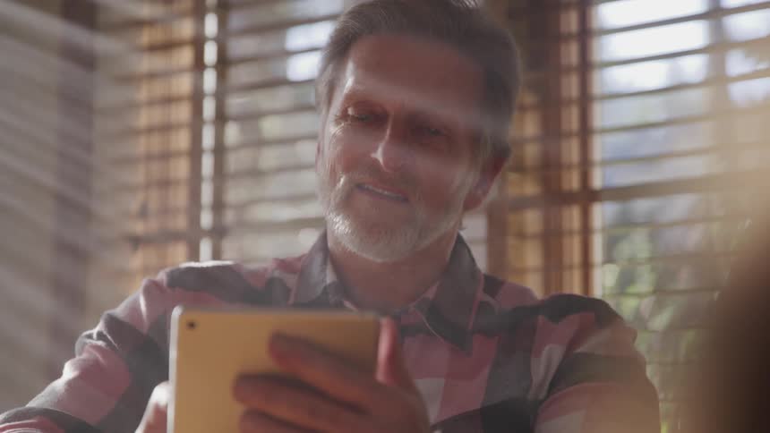 Middle aged man with a grey beard and a plaid shirt uses a tablet in a room