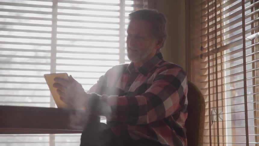 Bearded man uses a tablet sitting on a table in a room at home