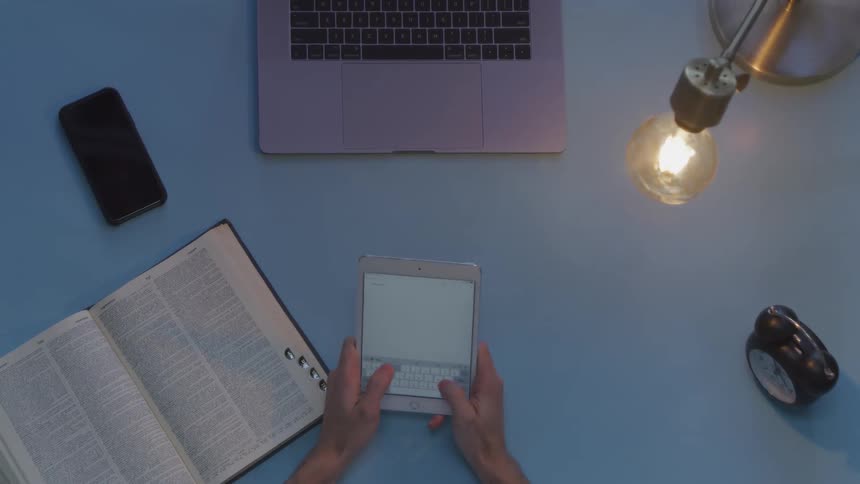 A male is quickly typing a word into their tablet device after reading it in a nearby dictionary