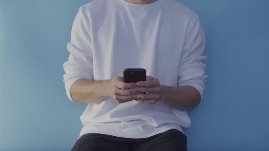 Man with a white sweatshirt and pants leans against a blue wall and sends someone a text message by smartphone