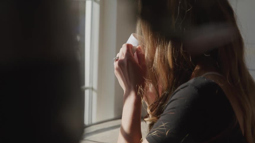 A woman sits by a window and takes a sip from a coffee mug