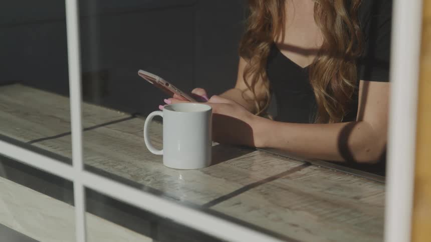 Female sitting at counter in a shop in front of the window with a coffee mug while typing on her cell phone