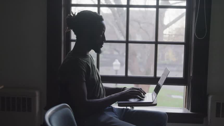 A man sits at a desk in a classroom and uses his laptop
