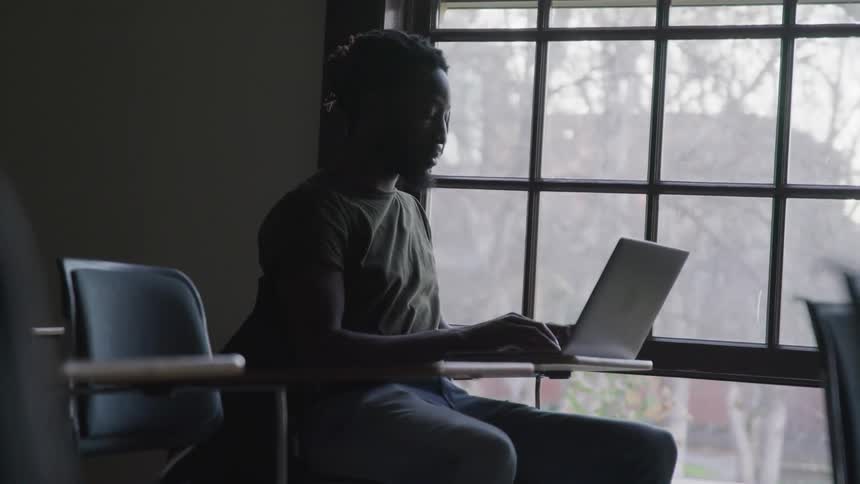 A student sits in a desk near a large window in a university classroom and types on his computer