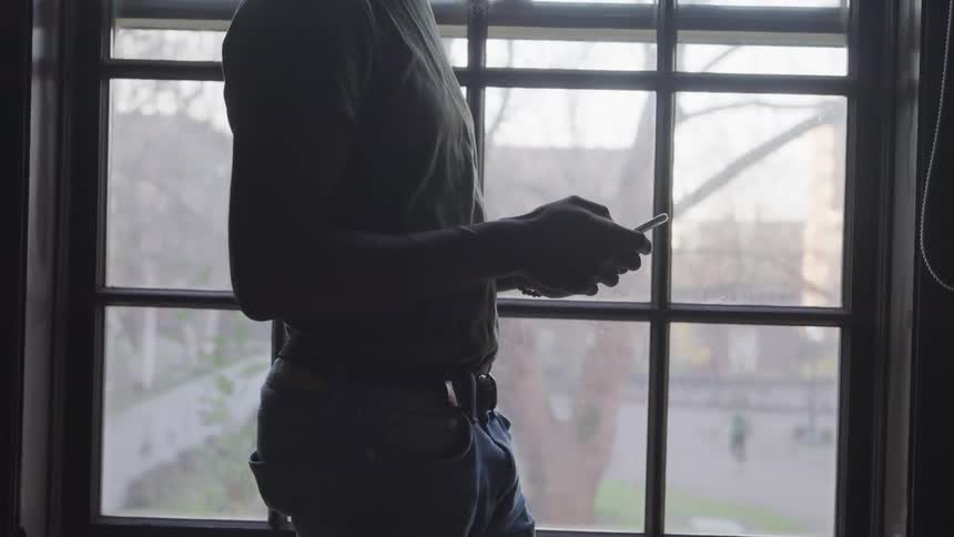 A person stands by a window scrolling on their cell phone