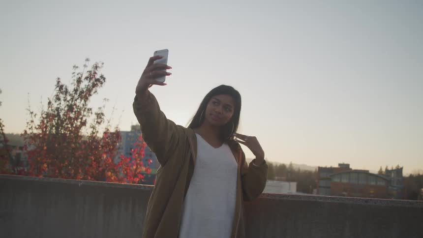 A woman taking pictures with a smart phone with trees the sky and buildings in the background