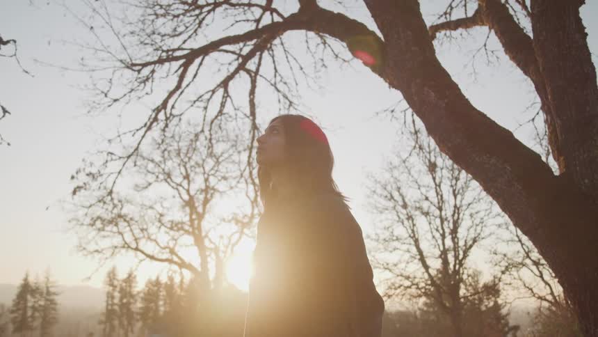Woman with straight hair and a green loose sweater top looks up at sky under a bare tree at sunset
