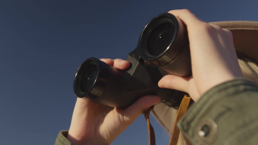 A woman looks around the area using a pair of binoculars while standing outdoors under a clear daytime sky