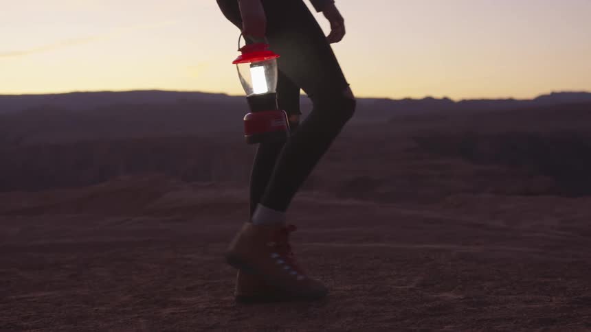 A woman holds a red lamp in her right hand as she hikes alone through the desert at sunset