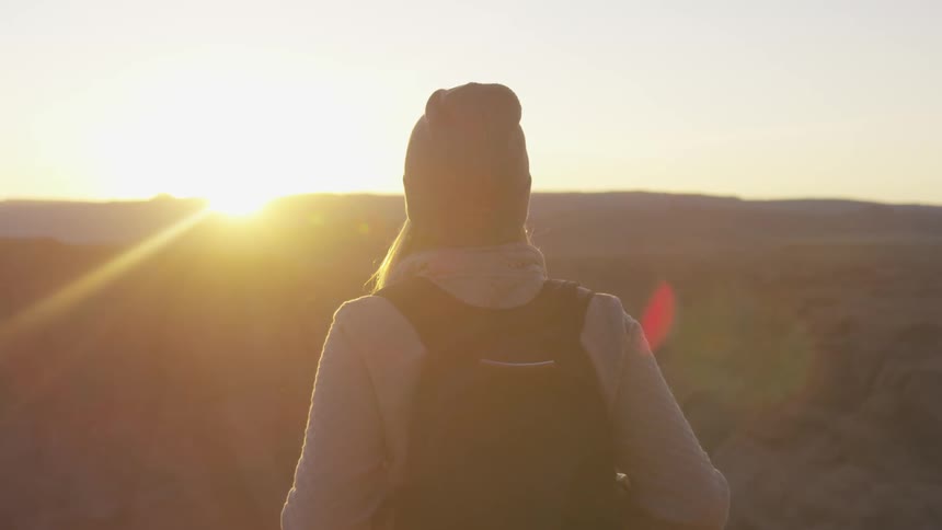 Woman with a beanie and a black backpack watches the sun setting on the horizon