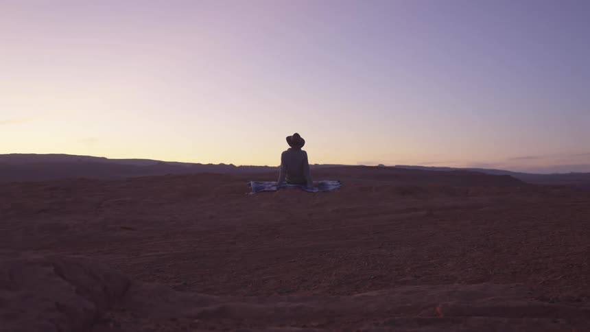 Woman wearing a hat sits on a blanket in the ground on the hills of a desert at sunset