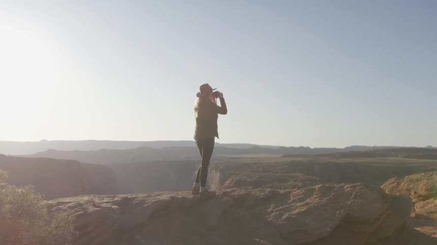 A woman stands on top of a tall mountain and uses her binoculars to see far in the distance