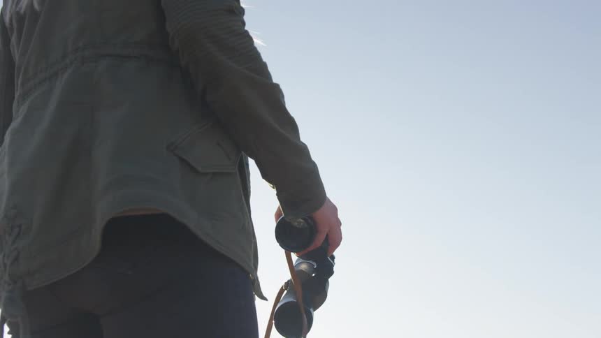 Young blonde woman walks to the edge of a cliff to use binoculars at sunset