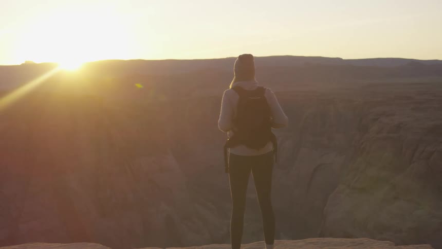 Woman stands looking down at canyon as sun sits on horizon