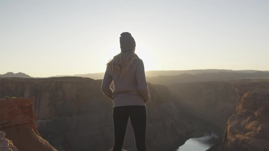 A woman stands on the cliff of a canyon and looks out over the landscape below as the sun peaks over the horizon