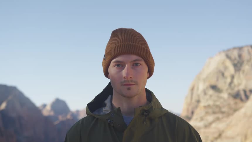 A man with a jacket and a hat standing and staring with a mountain range and the sky in the background