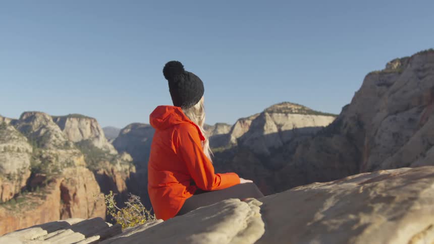 Woman wearing red jacket and black beanie sits on the rocks atop a mountain