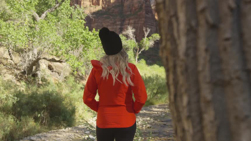 Young blonde woman wearing a red jacket and black beanie stands pensively on a trail through a canyon