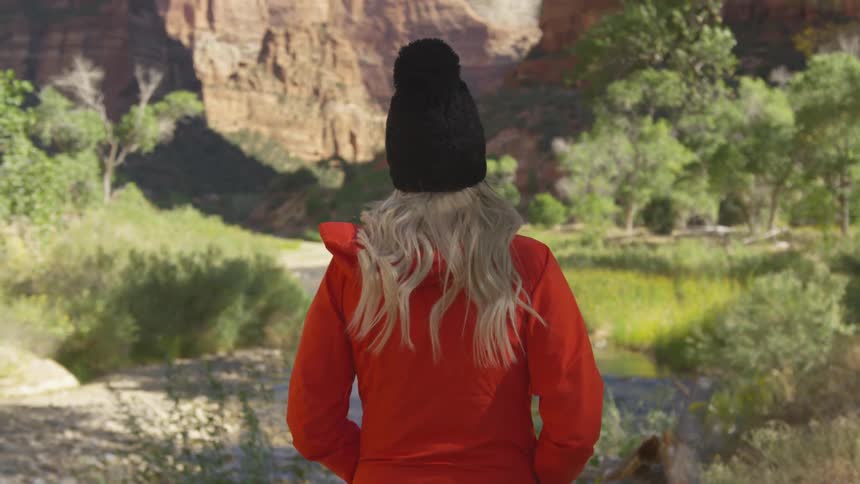 A woman in a red coat hikes along a trail at the base of a canyon