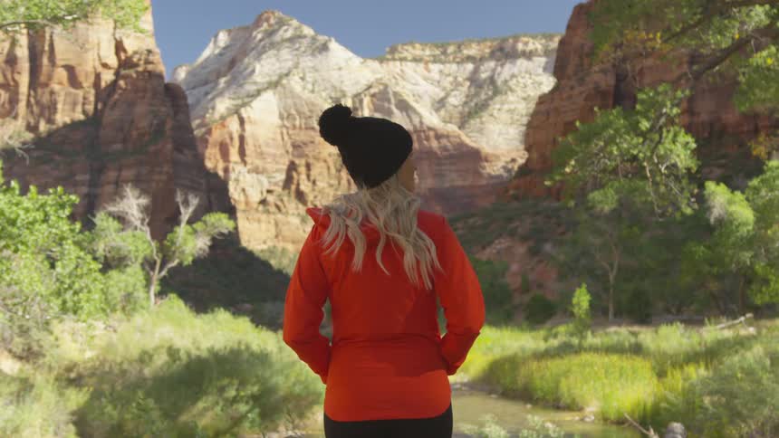 Blonde woman wearing red jacket and black beanie stands on the green vegetation at the mouth of a canyon