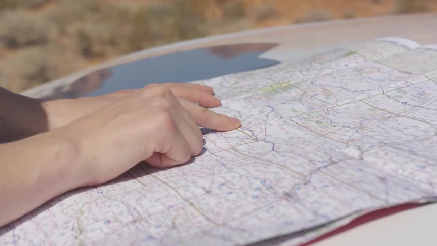 A woman runs her finger along a route on a paper map spread across the hood of her white car